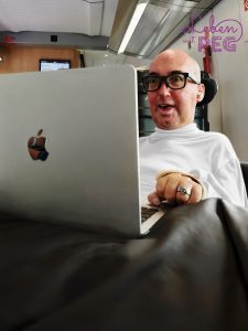 Bastian sitzt gut gelaunt mit seinem Laptop im Zug und arbeitet an seinen Projekten. Oben rechts das Logo des Blogs „Leben mit PEG“.